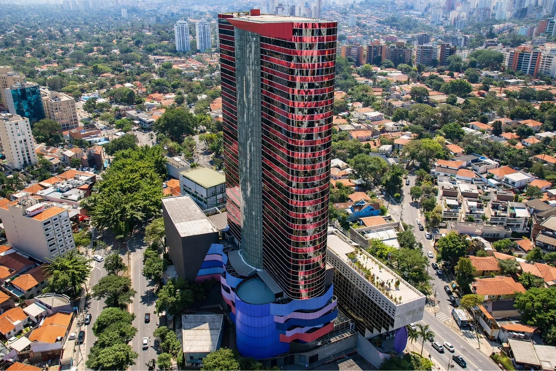 Aerial view of a modern red-and-glass high-rise tower at Pinheiros surrounded by a leafy residential neighborhood