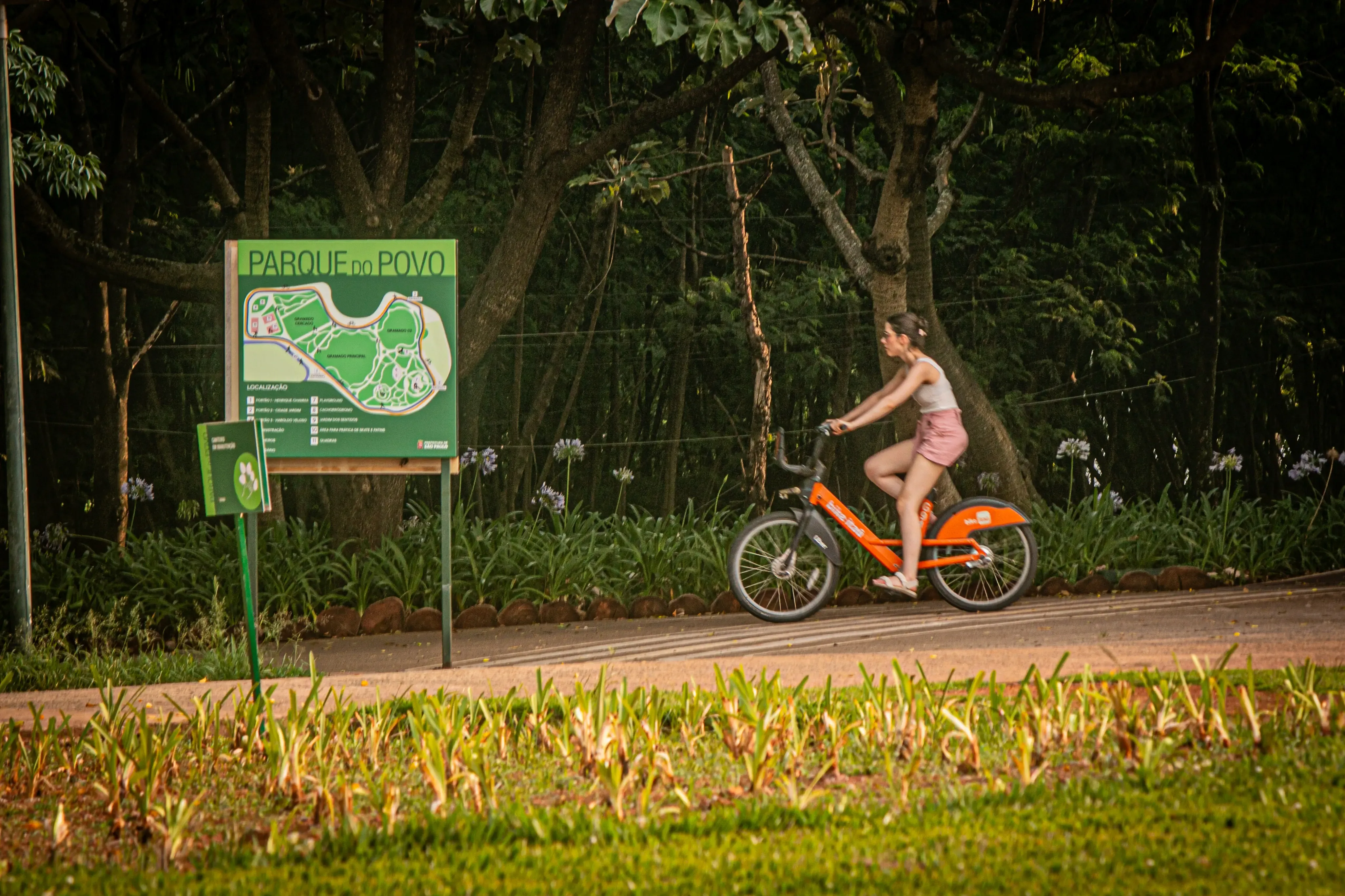 Woman riding an orange bike past the Parque do Povo sign and dense trees